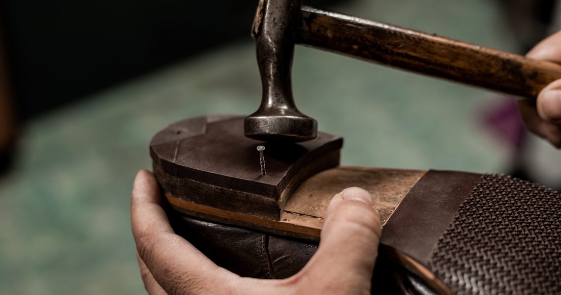 Male cobbler holding shoe and nailing a heel with hummer. Close-up on hands of professional shoemaker.
