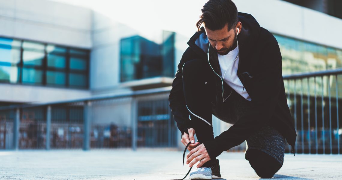 Young 20s male jogger in sports active wear and shoes preparing for cardio workout on street checking laces, male athlete tying sneakers before jogging dressed in trendy tracking clothes