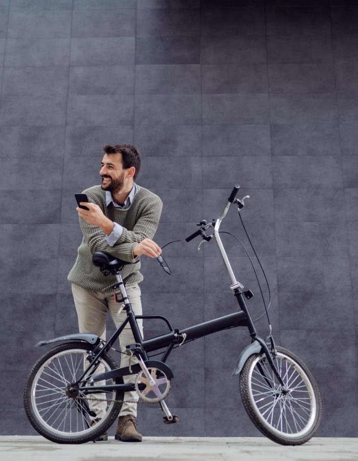 Full length of smiling handsome caucasian fashionable man leaning on his bicycle and holding smart phone and glasses. In background is gray wall.