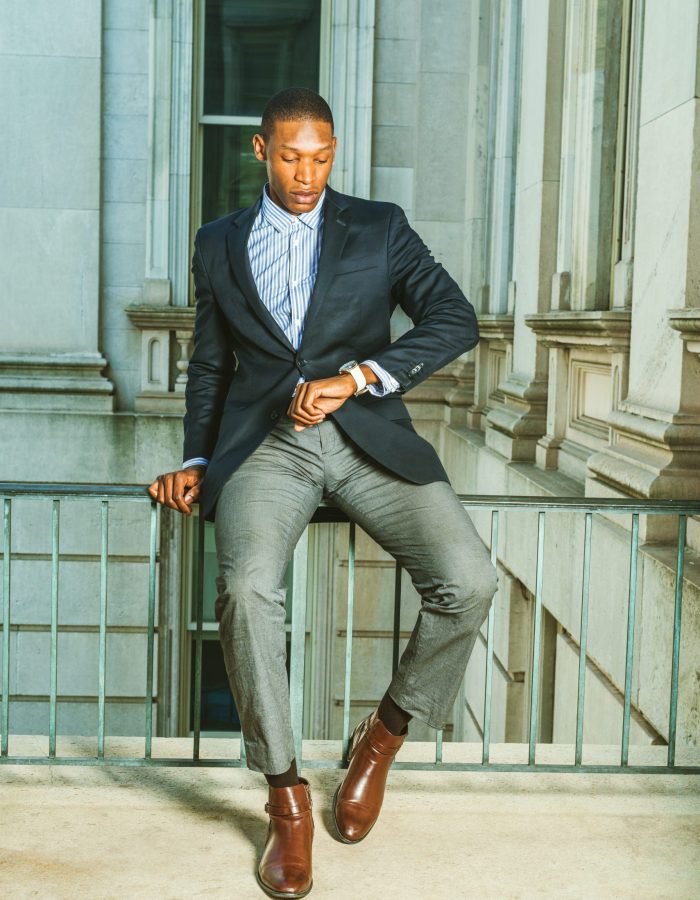 Man waiting for you. Dressing in black blazer, gray pants, brown leather shoes,  African American businessman sitting on railing in vintage style office, looking at his wristwatch. Time is money.